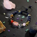 Beaded bracelet with dark blue and multicolored stones laid in a circle on a black surface, with crystals and chips around it; a pink rectangular charm sits at the front.
